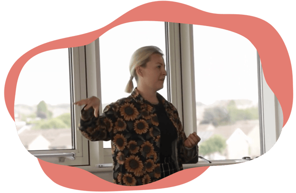 A person standing and speaking in front of large windows, gesturing with one hand during a presentation. A person standing and speaking in front of large windows, gesturing with one hand during a presentation.