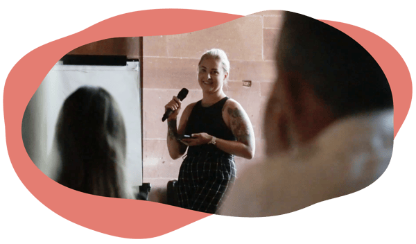 A person holding a microphone and smiling while speaking to a seated audience in a room with stone walls. A person holding a microphone and smiling while speaking to a seated audience in a room with stone walls.