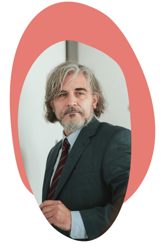 A person standing indoors, wearing a suit and looking thoughtfully to one side.