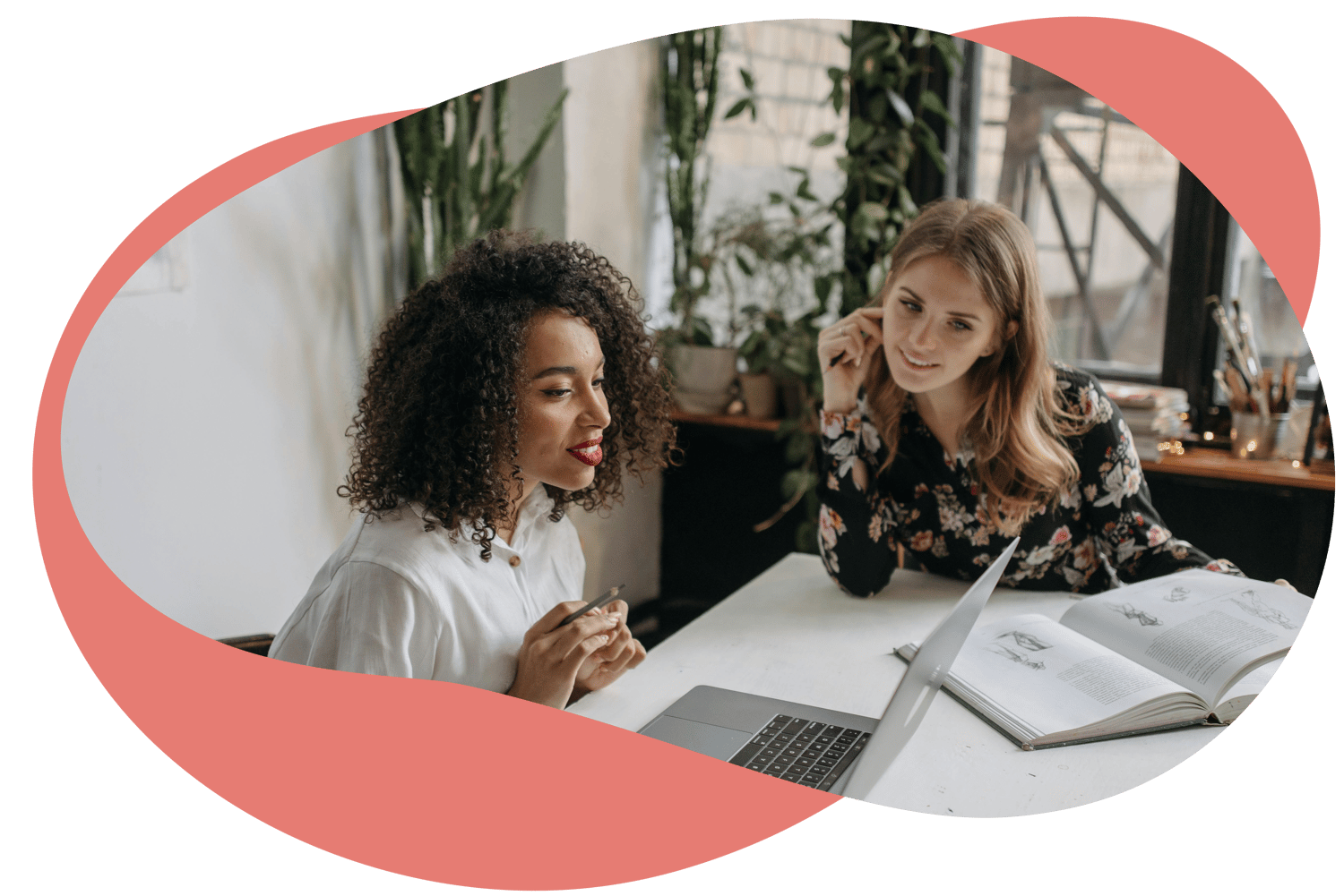 Two colleagues in a bright workspace discussing work together at a desk with a laptop and notebook.