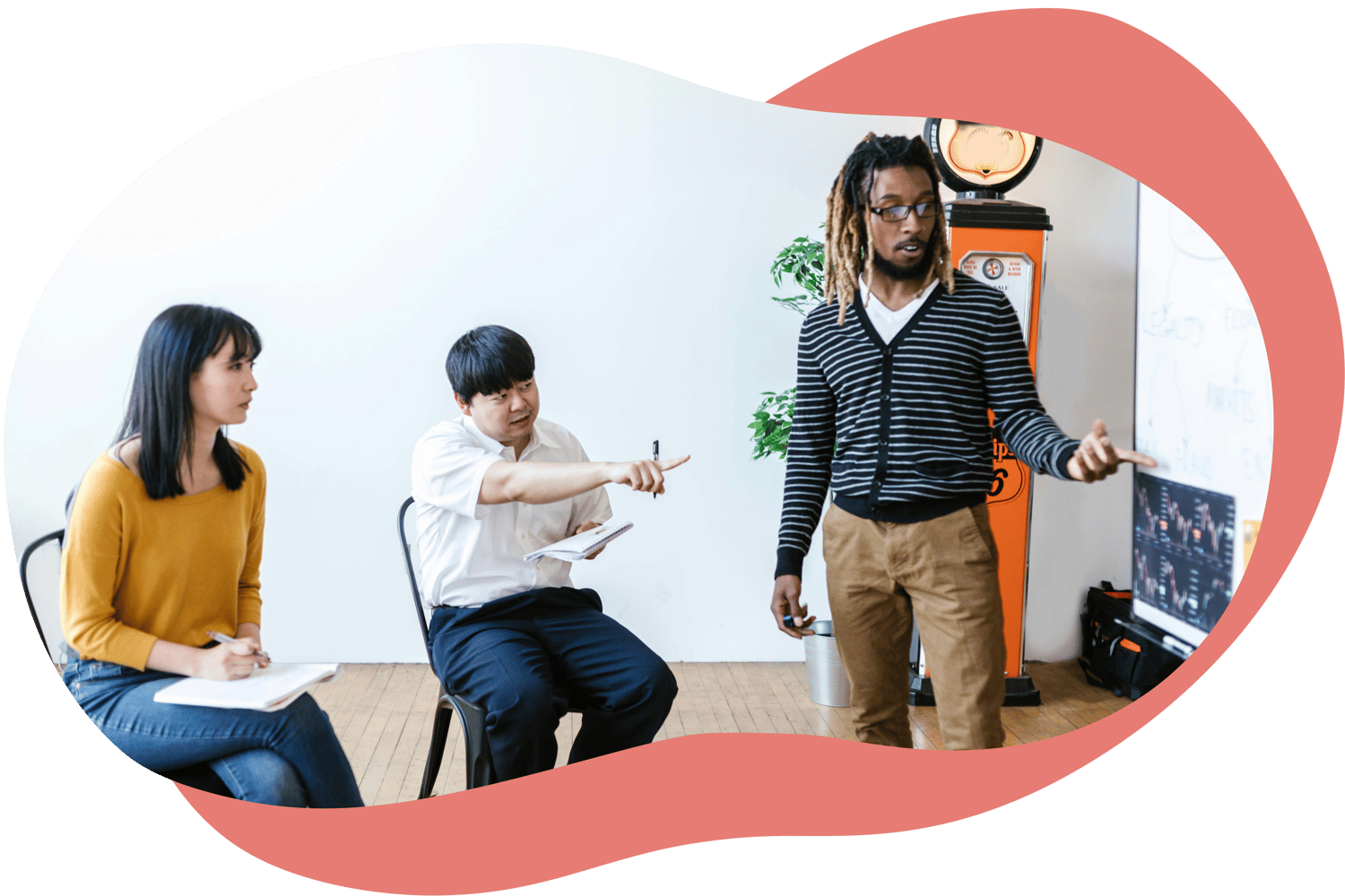 Three colleagues in a workshop setting, one person standing and explaining while others listen and take notes.