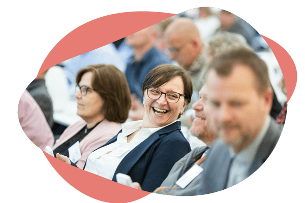 People seated at an event or conference, with one person in the centre smiling and engaging with others around them. People seated at an event or conference, with one person in the centre smiling and engaging with others around them.