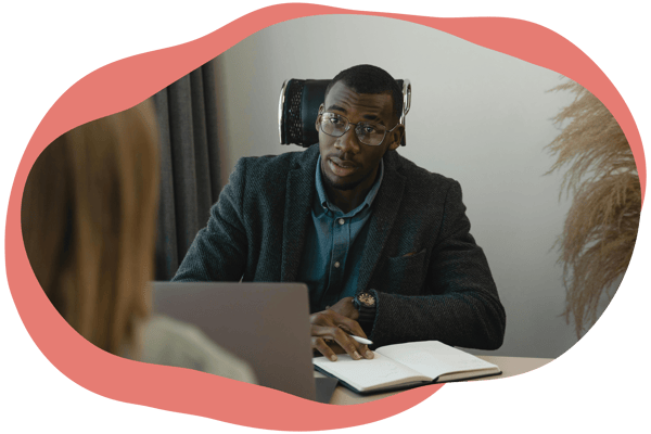 A person sitting at a desk with a notebook and laptop, speaking to someone seated opposite them. A person sitting at a desk with a notebook and laptop, speaking to someone seated opposite them.